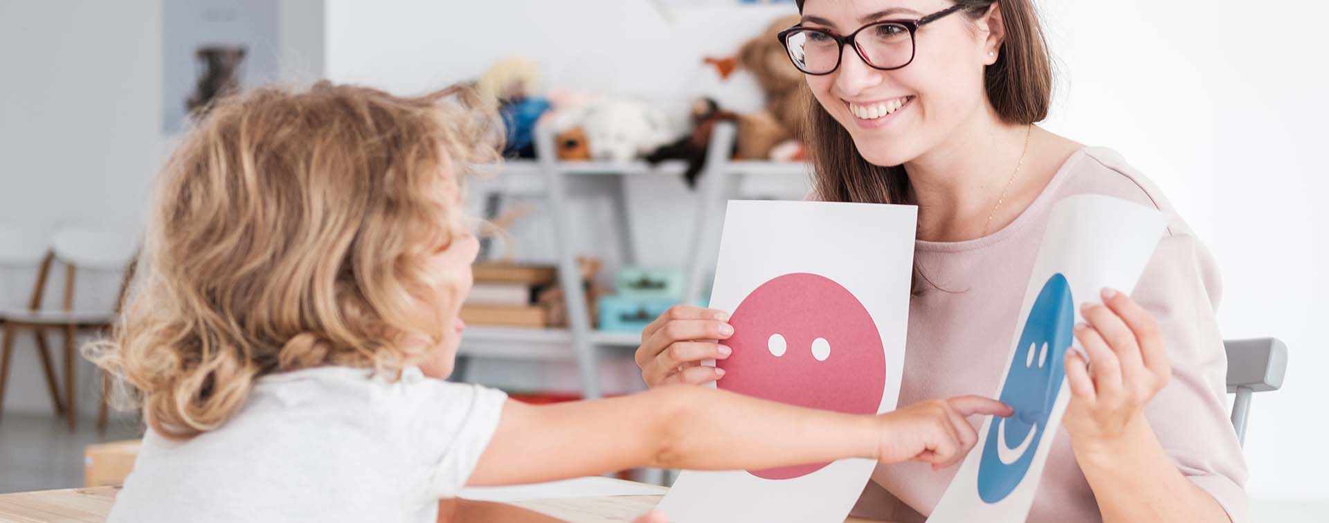 A child sitting across their teacher points to one of two smiley face images that she is holding up