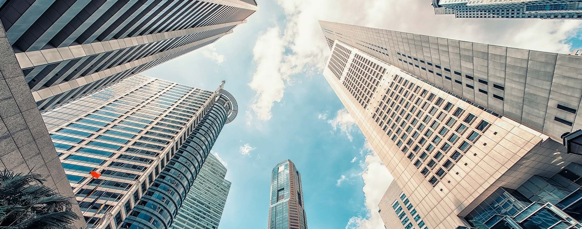 Several towering city buildings in Singapore seen from ground level looking up, with the blue sky and clouds above