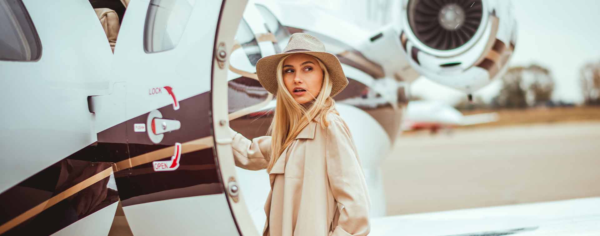 Young wealthy blonde female wearing beige hat and coat looking over her shoulder while embarking on a private airplane on a runway