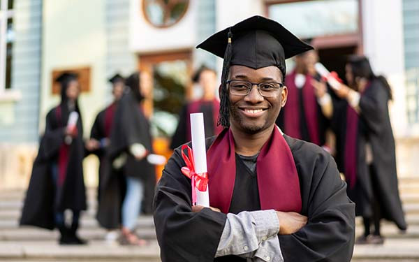 A smiling graduate holding his diploma after his graduation, standing on the university steps wearing his cap and gown