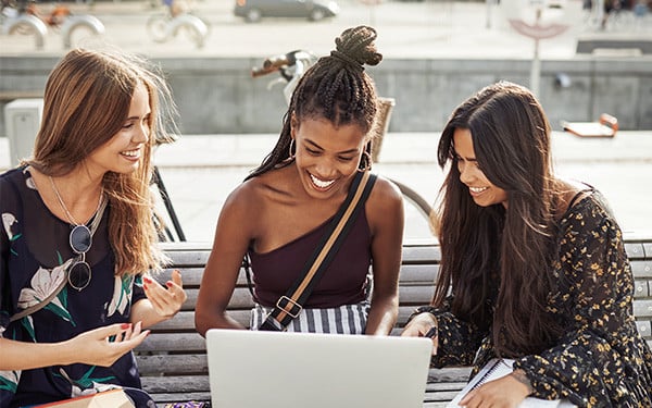 Three young female university students smiling while looking at a laptop on a bench outside